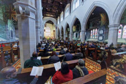 ACC delegates sit in a 17th century gothic cathedral with a colonnade of stone arches wooden pews, and high vaulted wooden ceiling