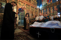A clergyman in cassock stands in a highly gilded icon lined church as people gather round a deceased person under a white pall