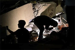 Two people crouch on rubble from a collapsed building